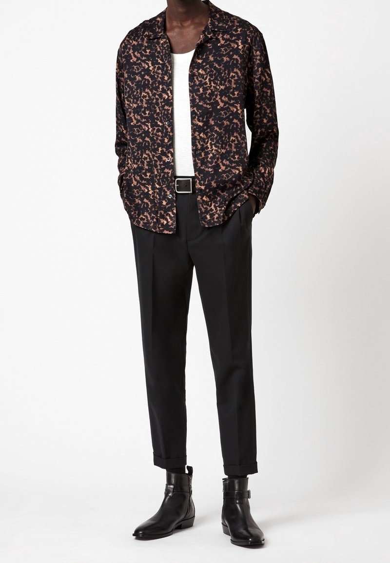 Camicia a maniche lunghe a fantasia nera e marrone indossata sopra una T-shirt bianca, abbinata a pantaloni neri su misura e stivaletti neri eleganti.