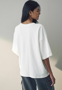 Oversized hvid t-shirt med faldne skuldre, blødt bomuldsstof, rund halsudskæring og afslappet pasform. Bæret med grå cargo-bukser.
