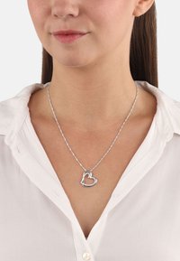 Silverfärgad hjärtformad halsband med en snodd kedja och en glittrande detalj, bärs över en vit skjorta med krage.