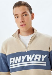 Sweatshirt zip-up beige et bleu avec un grand texte blanc "ANYWAY", comportant une petite fermeture éclair et des blocs de couleur horizontaux contrastés.