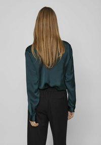 Blouse verte en satin à manches longues avec une texture lisse, coupe décontractée et poignets à boutons, associée à un pantalon noir taille haute doté de poches latérales.