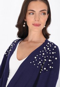 Cardigan marin en texture côtelée avec des embellissements en perles sur les épaules. Porté par-dessus un haut blanc avec un décolleté en V.