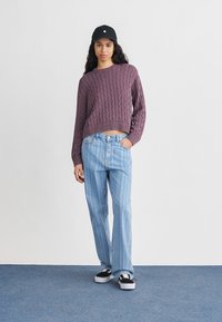 Γυναίκα στέκεται φορώντας μοβ πουλόβερ με πλέξη καλώδιο, ανοιχτό μπλε τζιν με ρίγες, μαύρο καπέλο και ασπρόμαυρα αθλητικά παπούτσια σε μπλε πάτωμα.