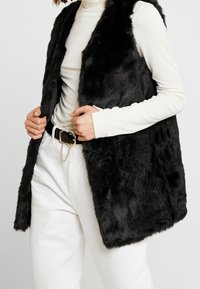 Svart konstläderväst med öppet framdesign, strukturerad yta, bärs över en vit långärmad skjorta, tillsammans med vita byxor och ett bälte.