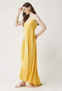 Maxikleid aus leuchtend gelbem Stoff mit Spaghettiträgern, ausgestattet mit einer Knopfleiste und Blumenstickerei entlang dem Saum. Flache Sandalen getragen.