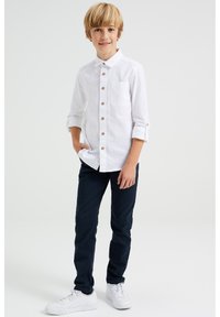 Bílá košile na knoflíky s dřevěnými knoflíky a náprsní kapsou, v kombinaci s tmavě modrými kalhotami a bílými teniskami. Volný střih, dlouhé rukávy posunuté nahoru.