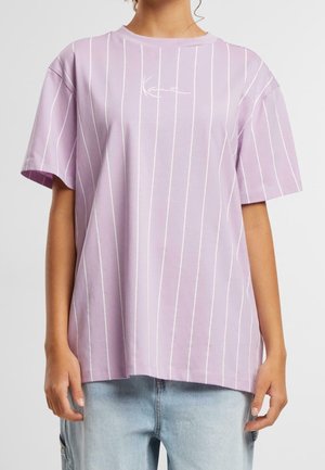 Person iført en løs, pastel lilla t-shirt med tynde hvide lodrette striber og casual lyseblå jeans, stående med afslappede arme.