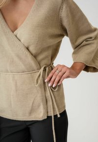 Beige Wickelsweater aus weichem Strickmaterial, mit V-Ausschnitt, 3/4-Ärmeln, gebundenem Taillenband und sichtbarer Textur an den Bündchen und am Saum.