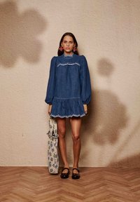 Τζιν φόρεμα με φουσκωτά μανίκια και κεντητές λεπτομέρειες, με φαρδιά κάτω ποδόγυρο, σετ με τσάντα ώμου με λουλουδάτο σχέδιο και σκούρα παπούτσια.