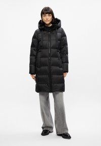 Cappotto imbottito nero con cappuccio, dotato di zip frontale, cinghie regolabili e motivo trapuntato. Abbinato a pantaloni grigi a gamba larga.