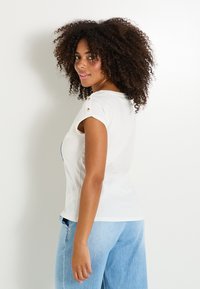 Camiseta blanca de manga corta con detalles de botones en los hombros, combinada con pantalones de mezclilla azul claro. Tela de algodón suave, corte relajado.