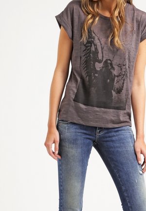 Woman wearing faded black graphic t-shirt with angel wings design and blue faded jeans standing against a white background.