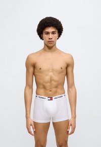 Άσπρα βαμβακερά μπόξερ με μαύρη μέση που φέρει το λογότυπο "TOMMY HILFIGER". Λείο ύφασμα και εφαρμοστό σχέδιο. Το μοντέλο στέκεται όρθιο.