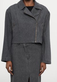 Giacca di denim grigia dal design corto, con ampio colletto, chiusura con cerniera angolata e tasche laterali con zip. Tessuto liscio, cuciture minimali.