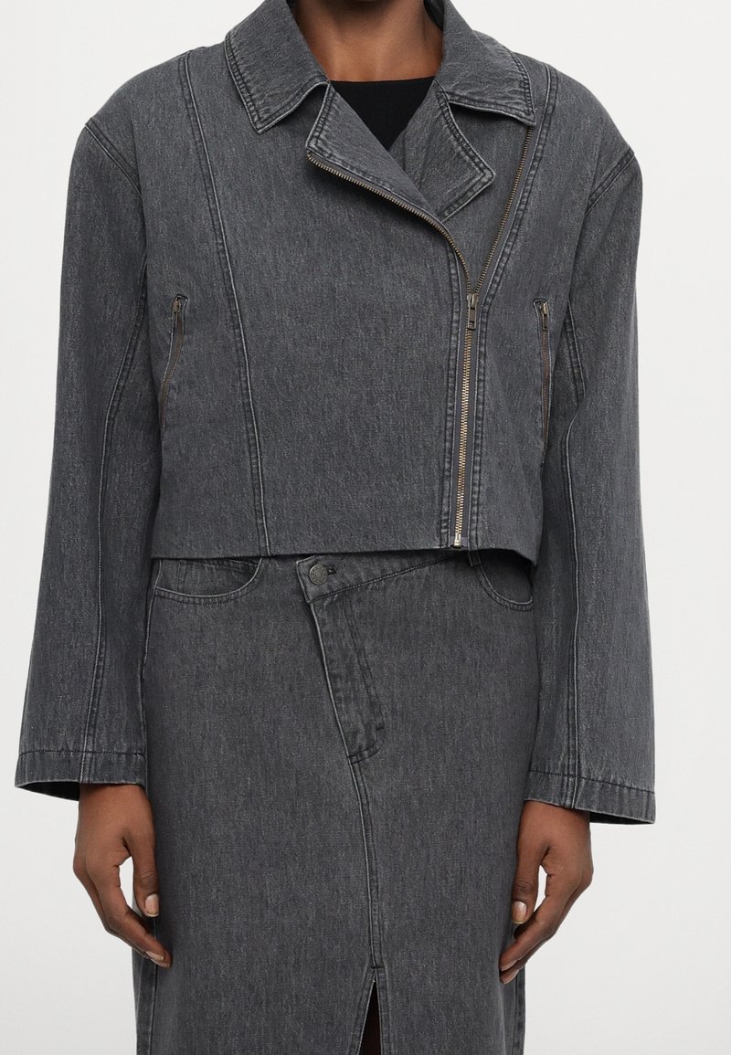 Giacca di denim grigia dal design corto, con ampio colletto, chiusura con cerniera angolata e tasche laterali con zip. Tessuto liscio, cuciture minimali.