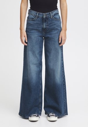 Vaqueros de mezclilla azul de pierna ancha con lavado desgastado, combinados con top negro y zapatillas negras y blancas sobre fondo blanco.