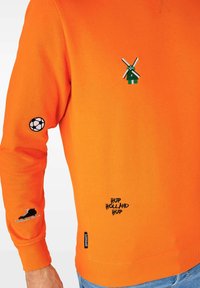 Oranžinė džemperis su siuvinėtais raštais: žalia vėjo malūnas, futbolo kamuolys, ledo pačiūžos ir tekstas "HUP HOLLAND HUP". Medvilninė medžiaga, apykaklė su apvaliu kaklu.