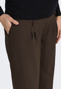 Braune Hose aus glattem Stoff, mit seitlichen Taschen, geradem Schnitt und dezenten Falten am Bund.