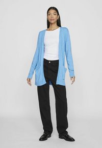 Heledad sinine, kudum kaartigan avatud esiosa, pikkade varrukatega ja kahe ees oleva taskuga, kombineeritud mustade laiemate pükstega ja mustade kingadega.