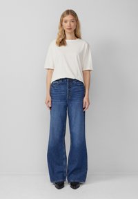 Losse blauwe jeans met een hoge taille, met een knoopsluiting aan de voorkant en wijde pijpen. Gecombineerd met een eenvoudig wit T-shirt met korte mouwen.