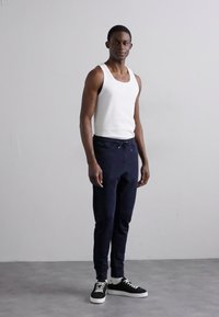 Λευκή αμάνικη μπλούζα, ναυτικές φόρμες με κορδόνι, μαύρα αθλητικά παπούτσια με λευκή σόλα, στέκεται σε ουδέτερο φόντο.