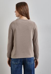 Beige pitkähihainen toppi pyöreällä kaula-aukolla ja ribbikuvioinnilla, yhdistettynä sinisiin leveälahkeisiin farkkuihin, näkyy takaa.