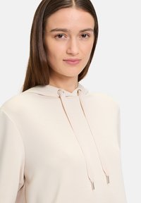 Beżowa bluza z kapturem na sznurki, z metalowymi elementami w kolorze srebrnym, miękka w dotyku i luźna, podkreślająca minimalistyczny design.