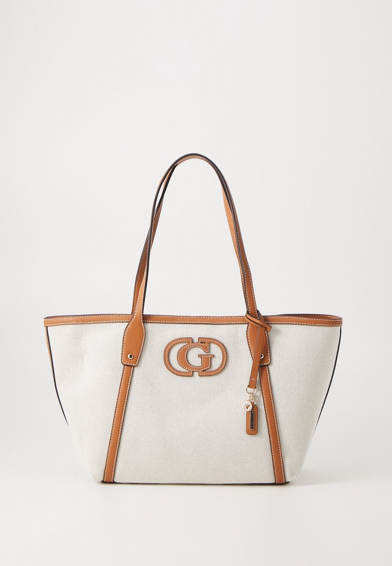 Sac fourre-tout en toile beige avec garniture en cuir marron clair, poignées doubles, patch logo décoratif et un petit charme suspendu sur le côté avant droit.