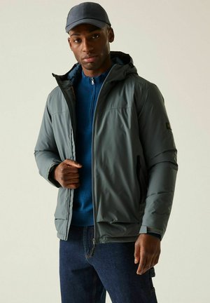 Mężczyzna ubrany w szarą kurtkę z kapturem na niebieskim swetrze z zamkiem do połowy długości, ciemne jeansy i szara czapka, stojący na jednolitym tle.