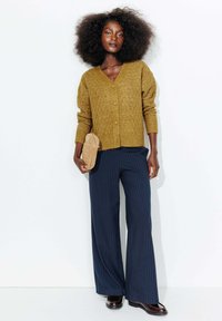 Cardigan en laine jaune moutarde avec un motif en losange, fermeture à boutons, associé à un pantalon large à rayures bleu marine et des chaussures marron.