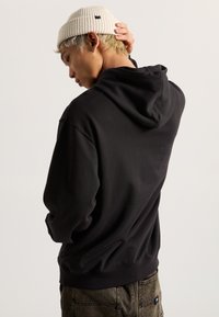 Sudadera con capucha negra de textura suave, con puños acanalados. El modelo lleva un gorro crema. Las costuras visibles acentúan el diseño.