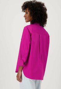 Heldere fuchsia langemouwen shirt met een relaxte pasvorm, geknoopte manchetten en een rugplooidetail, gecombineerd met lichtblauwe jeans.