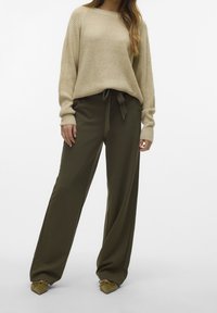 Maglione beige lavorato a maglia con maniche lunghe, abbinato a pantaloni larghi color oliva con cintura annodata. Scarpe color oliva completano l'outfit.