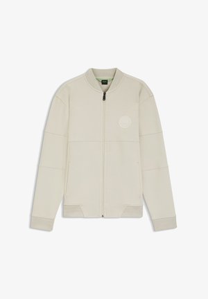 Vaaleanbeige vetoketjullinen takki pehmeästä kankaasta, jossa pyöreä rintalogo, resorit hihansuissa ja paneelirakenteinen muotoilu.