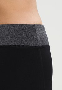 Černé legíny s šedým elastickým pasem. Látka je hladká a přiléhavá, s viditelnými švy podél švů.