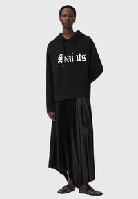 Felpa nera oversize con la scritta "Saints" in bianco, abbinata a una gonna nera plissettata lunga fino al pavimento e calzature nere con borchie.