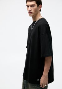 Tricou negru supradimensionat din bumbac, cu mâneci scurte și guler rotund. Detaliu notabil: logo mic la tiv.