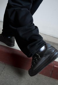 Černé tenisky s robustním designem, vyrobené z texturovaného materiálu, s výraznými tkaničkami a silnou podrážkou, v kombinaci s volnými černými kalhotami.