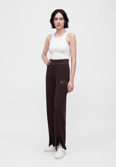 Femme aux cheveux courts et foncés portant un haut blanc sans manches, un pantalon large foncé avec des fentes et le logo "A|X", ainsi que des baskets blanches, debout sur un fond uni.