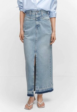 Femme portant une jupe maxi en denim bleu clair avec une fente à l'avant, ourlet effiloché, associée à une chemise bleu clair aux manches retroussées et des sandales blanches à bout carré.