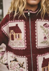 Bordeauxroter Strickpullover mit Reißverschluss, mit gemusterten Quadraten, die ein Haus und florale Designs in sanften Pastelltönen zeigen.