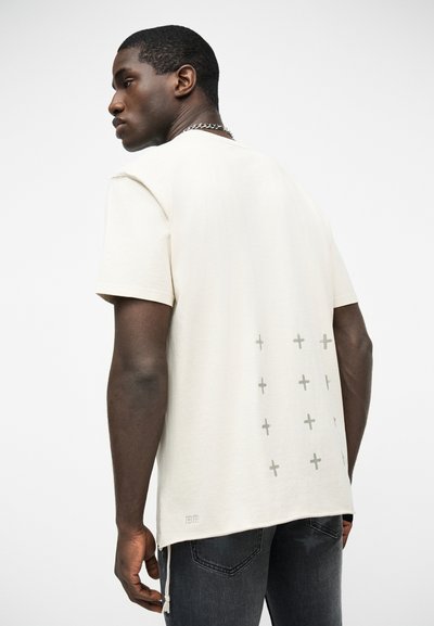 Άνδρας που φορά εκρού κοντομάνικο πουκάμισο με σχέδια σταυρών στην πλάτη και σκούρο τζιν, γυρισμένος πλάγια με το κεφάλι στραμμένο προς το πλάι.