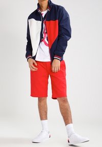 Marinblå och röd bomberjacka med vita paneler, röda shorts och vita sneakers. Har dragkedjestängning och randiga detaljer.