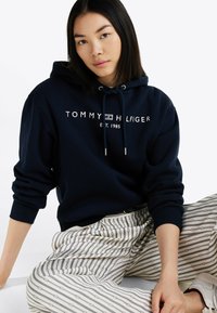 Granatowy hoodie z sznurkiem i białym logo, połączony z beżowymi spodniami w paski z geometrycznym wzorem. Miękka tkanina i luźny krój.