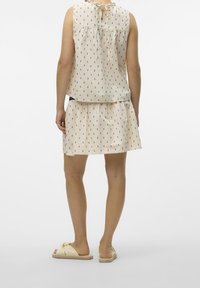 Robe de couleur crème avec taille froncée, présentant de petits motifs noirs. L'encolure au dos a une boucle avec un nœud. Assortie avec des sandales de couleur claire.