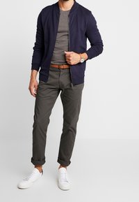 Mężczyzna w granatowej bluzie na zamek, szarym podkoszulku, ciemnych spodni z podwiniętymi nogawkami, brązowym pasku, zegarku na nadgarstku i białych trampkach stoi na tle białego tła.