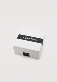 Boîte carrée blanche avec une bande noire en haut affichant la marque "Calvin Klein", posée sur une surface claire.