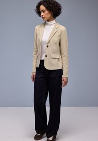 Blazer beige texturé avec une coupe ajustée, fermeture à un bouton et deux poches avant, associé à un jean large bleu foncé et des baskets marron.