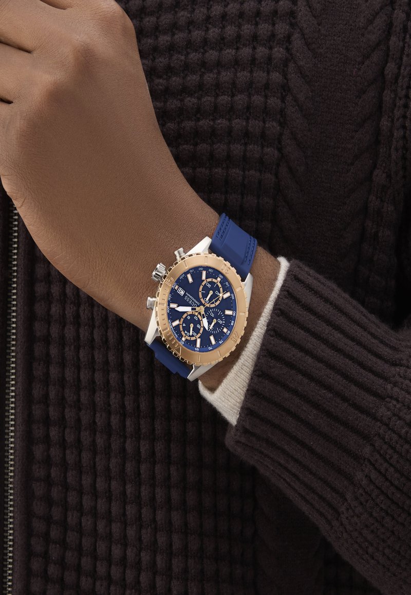 Reloj cronógrafo azul con acentos dorados, correa de goma azul texturizada y un dial azul detallado. Lleva puesto sobre la manga de un suéter de punto marrón.