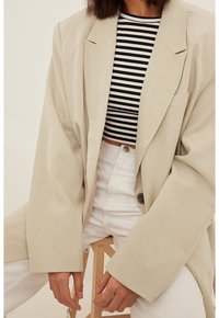 Beige oversized blazer met grote revers, gedragen over een zwart-wit gestreepte cropped top, gecombineerd met witte broek met hoge taille.